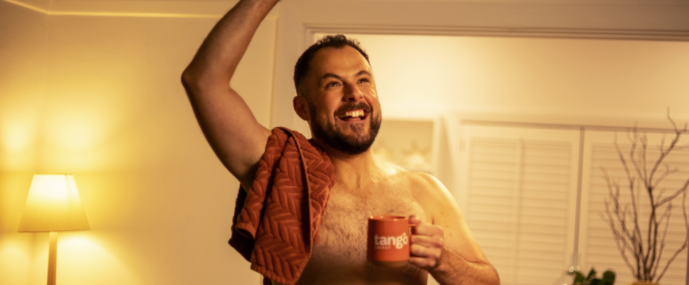 <div>Energy that's stripped right back.</div>
A man smiling and looking up. He is shirtless and is standing in front of a basket of laundry. One hand is in the air and one hand is holding a mug.